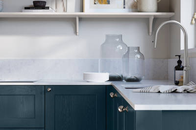 Ludlow, a traditional Shaker-style premium feel kitchen range with a visible woodgrain on its panelled doors here shown in the shade Voyage Blue styled with steel knobs, steel tap, a white marbled worktop with matching splashback.