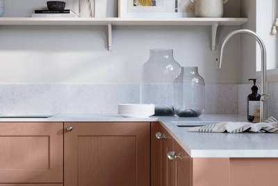 Ludlow, a traditional Shaker-style premium feel kitchen range with a visible woodgrain on its panelled doors here shown in the shade Clay styled with steel knobs, steel tap, a white marbled worktop with matching splashback.