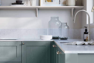 Ludlow, a traditional Shaker-style premium feel kitchen range with a visible woodgrain on its panelled doors here shown in the light shade Coastland styled with steel knobs, steel tap, a white marbled worktop with matching splashback.