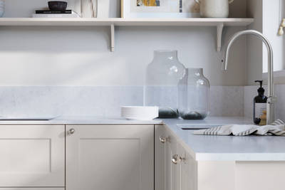Ludlow, a traditional Shaker-style premium feel kitchen range with a visible woodgrain on its panelled doors here shown in the light shade Fresh Linen styled with steel knobs, steel tap, a white marbled worktop with matching splashback.