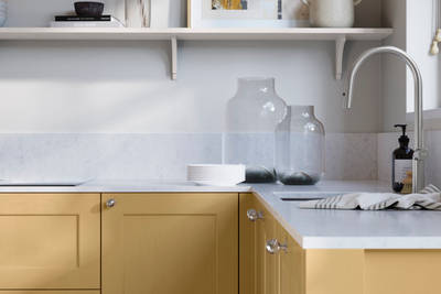 Ludlow, a traditional Shaker-style premium feel kitchen range with a visible woodgrain on its panelled doors here shown in the yellow shade Harvest styled with steel knobs, steel tap, a white marbled worktop with matching splashback.