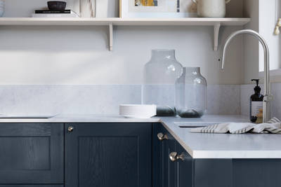 Ludlow, a traditional Shaker-style premium feel kitchen range with a visible woodgrain on its panelled doors here shown in the dark blue shade Midnight styled with steel knobs, steel tap, a white marbled worktop with matching splashback.