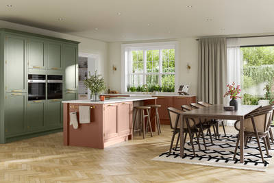 Open floorplan kitchen and dining rool with tall Wardley kitchen cabinetry from Magnet with built-in ovens and a kitchen island, in the shades Thyme and Clay, with a large wooden dining room table.