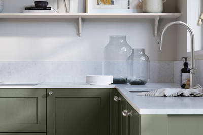 Wardley Thyme kitchen cabinetry with steel knobs, steel tap, white marbled worktop and matching backsplash. An open shelf on the wall above cabinets with ceramics and a vase of flowers.