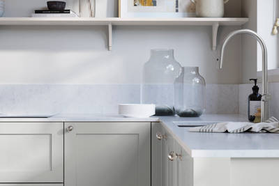 Wardley Washed Cotton kitchen cabinetry with steel knobs, steel tap, white marbled worktop and matching backsplash. An open shelf on the wall above cabinets with ceramics and a vase of flowers.