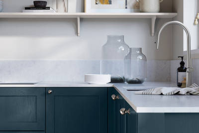 Wardley Voyage Blue kitchen cabinetry with steel knobs, steel tap, white marbled worktop and matching backsplash. An open shelf on the wall above cabinets with ceramics and a vase of flowers.