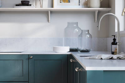 Wardley Carrington Blue kitchen cabinetry with steel knobs, steel tap, white marbled worktop and matching backsplash. An open shelf on the wall above cabinets with ceramics and a vase of flowers.