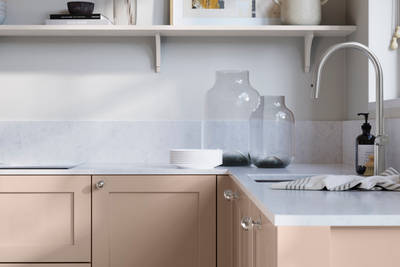 Wardley Chalk Blush kitchen cabinetry with steel knobs, steel tap, white marbled worktop and matching backsplash. An open shelf on the wall above cabinets with ceramics and a vase of flowers.