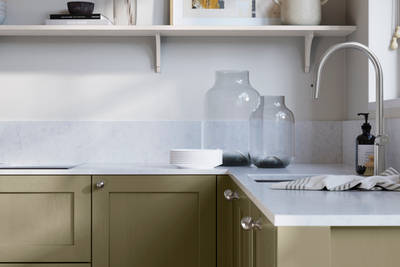 Wardley Balmoral kitchen cabinetry with steel knobs, steel tap, white marbled worktop and matching backsplash. An open shelf on the wall above cabinets with ceramics and a vase of flowers.