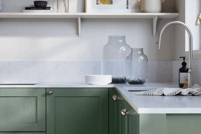 Wardley Eucalyptus kitchen cabinetry with steel knobs, steel tap, white marbled worktop and matching backsplash. An open shelf on the wall above cabinets with ceramics and a vase of flowers.