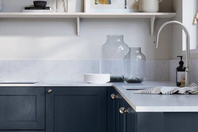 Wardley Midnight kitchen cabinetry with steel knobs, steel tap, white marbled worktop and matching backsplash. An open shelf on the wall above cabinets with ceramics and a vase of flowers.