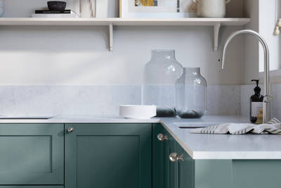 Wardley Morris Blue kitchen cabinetry with steel knobs, steel tap, white marbled worktop and matching backsplash. An open shelf on the wall above cabinets with ceramics and a vase of flowers.