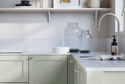Wardley Seagrass kitchen cabinetry with steel knobs, steel tap, white marbled worktop and matching backsplash. An open shelf on the wall above cabinets with ceramics and a vase of flowers.