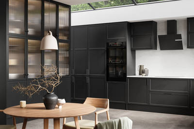 View of modern shaker kitchen Brunswick Sumi Black, with screen extractor over induction hob, tall fluted glass doorsvitrine cabinets, black frame skylights with wood dining table and chairs in foreground.