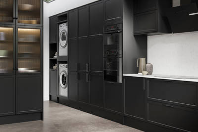 View of Modern Shaker Brunswick Sumi Black laundry room from kitchen, with integrated laundry machine and dryer as well as ovens in tall cabinetry, white worktop and splashback Silestone Miami Vena and vitrine cabinets with fluted glass doors.