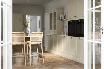 View from patio through opened white frame glass doors of traditional Shaker-style kitchen Ludlow Parchment with tactile woodgrain, tall cabinets, brass cup handles and knobs, integrated ovens, a kitchen island with rattan bar stools, a vase holding foliage, and light wood herringbone flooring.