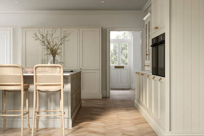 Traditional Shaker-style kitchen Ludlow Parchment with tactile woodgrain, tall cabinets, brass cup handles and knobs, integrated ovens, a kitchen island with rattan bar stools, a vase holding foliage, and light wood herringbone flooring.