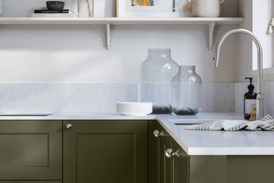 Ludlow, a traditional Shaker-style premium feel kitchen range with a visible woodgrain on its panelled doors here shown in the shade Tailored Green styled with steel knobs, steel tap, a white marbled worktop with matching splashback.