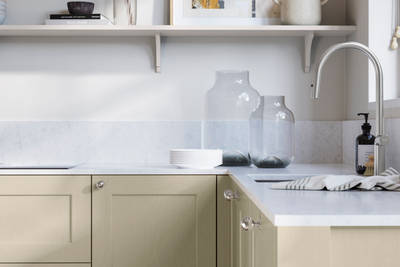 Ludlow, a traditional Shaker-style premium feel kitchen range with a visible woodgrain on its panelled doors here shown in the shade Parchment styled with steel knobs, steel tap, a white marbled worktop with matching splashback.