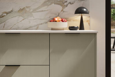 Fluted front kitchen Reed from Magnet in neutral shade French Clay, with a marbled splashback and black handles, herringbone wood flooring.