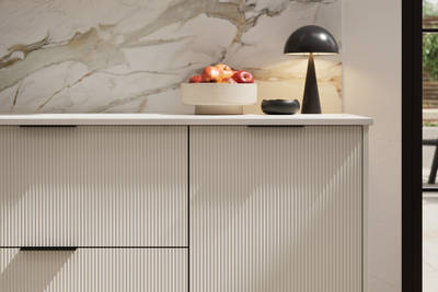 Fluted front kitchen Reed from Magnet in light neutral shade Limestone, with a marbled splashback and black handles, herringbone wood flooring.