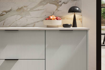 Fluted front kitchen Reed from Magnet in light neutral shade Washed cotton, with a marbled splashback and black handles, herringbone wood flooring.
