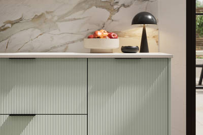 Fluted front kitchen Reed from Magnet in light green shade Seagrass, with a marbled splashback and black handles, herringbone wood flooring.