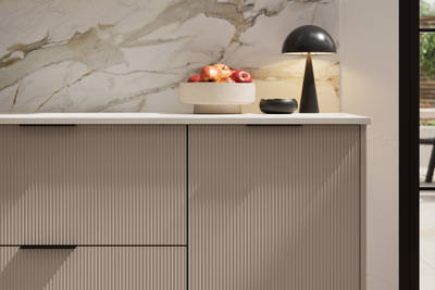 Fluted front kitchen Reed from Magnet in neutral shade Cornish Clay, with a marbled splashback and black handles, herringbone wood flooring.