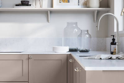 Wardley Cornish Clay kitchen cabinetry with steel knobs, steel tap, white marbled worktop and matching backsplas, an open shelf on the wall above cabinets with ceramics and a vase of flowers.