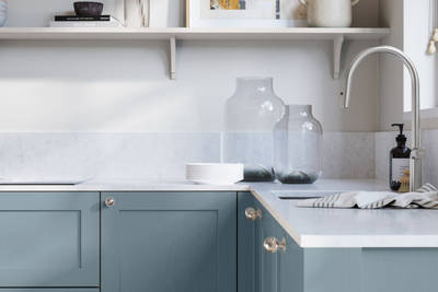 Wardley Portland Blue kitchen cabinetry with steel knobs, steel tap, white marbled worktop and matching backsplas, an open shelf on the wall above cabinets with ceramics and a vase of flowers.
