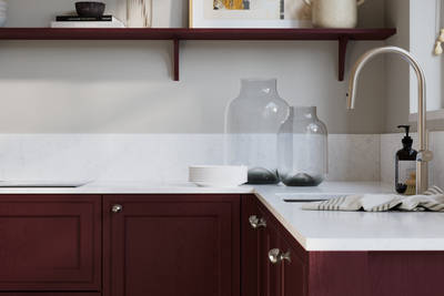 Bewdley Morello painted wood grain Shaker kitchen cabinetry with steel knobs, steel tap, white marbled worktop and matching backsplash, an open shelf on the wall above cabinets with ceramics and a vase of flowers.