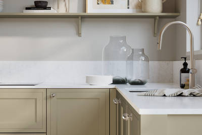 Bewdley Parchment painted wood grain Shaker kitchen cabinetry with steel knobs, steel tap, white marbled worktop and matching backsplash, an open shelf on the wall above cabinets with ceramics and a vase of flowers.