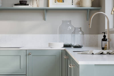 Bewdley Sky painted wood grain Shaker kitchen cabinetry with steel knobs, steel tap, white marbled worktop and matching backsplash, an open shelf on the wall above cabinets with ceramics and a vase of flowers.