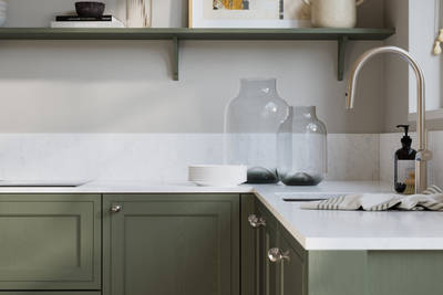 Bewdley Thyme painted wood grain Shaker kitchen cabinetry with steel knobs, steel tap, white marbled worktop and matching backsplash, an open shelf on the wall above cabinets with ceramics and a vase of flowers.