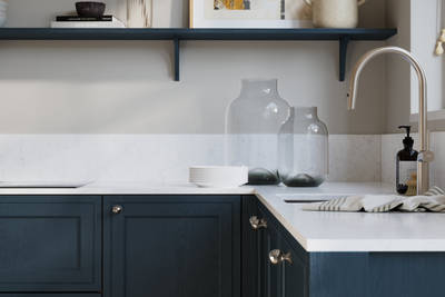 Bewdley Voyage Blue painted wood grain Shaker kitchen cabinetry with steel knobs, steel tap, white marbled worktop and matching backsplash, an open shelf on the wall above cabinets with ceramics and a vase of flowers.