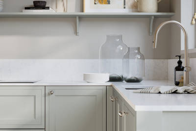 Bewdley Washed Cotton painted wood grain Shaker kitchen cabinetry with steel knobs, steel tap, white marbled worktop and matching backsplash, an open shelf on the wall above cabinets with ceramics and a vase of flowers.