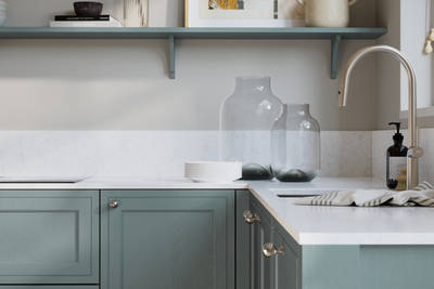 Bewdley Duck Egg painted wood grain Shaker kitchen cabinetry with steel knobs, steel tap, white marbled worktop and matching backsplash, an open shelf on the wall above cabinets with ceramics and a vase of flowers.