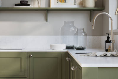 Bewdley Misty Moors painted wood grain Shaker kitchen cabinetry with steel knobs, steel tap, white marbled worktop and matching backsplash, an open shelf on the wall above cabinets with ceramics and a vase of flowers.