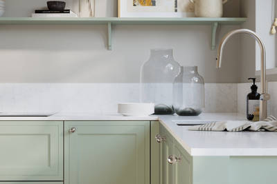 Bewdley Seagrass painted wood grain Shaker kitchen cabinetry with steel knobs, steel tap, white marbled worktop and matching backsplash, an open shelf on the wall above cabinets with ceramics and a vase of flowers.