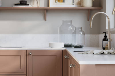 Bewdley Baked Clay painted wood grain Shaker kitchen cabinetry with steel knobs, steel tap, white marbled worktop and matching backsplash, an open shelf on the wall above cabinets with ceramics and a vase of flowers.