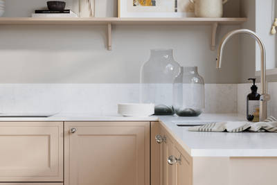 Bewdley Chalk Blush painted wood grain Shaker kitchen cabinetry with steel knobs, steel tap, white marbled worktop and matching backsplash, an open shelf on the wall above cabinets with ceramics and a vase of flowers.