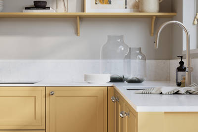 Bewdley Harvest painted wood grain Shaker kitchen cabinetry with steel knobs, steel tap, white marbled worktop and matching backsplash, an open shelf on the wall above cabinets with ceramics and a vase of flowers.