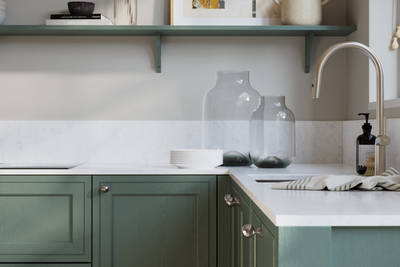 Bewdley Eucalyptus Green painted wood grain Shaker kitchen cabinetry with steel knobs, steel tap, white marbled worktop and matching backsplash, an open shelf on the wall above cabinets with ceramics and a vase of flowers.
