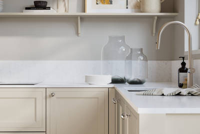 Bewdley Limestone painted wood grain Shaker kitchen cabinetry with steel knobs, steel tap, white marbled worktop and matching backsplash, an open shelf on the wall above cabinets with ceramics and a vase of flowers.