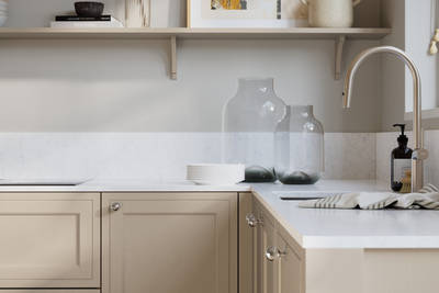 Bewdley Pebble Path painted wood grain Shaker kitchen cabinetry with steel knobs, steel tap, white marbled worktop and matching backsplash, an open shelf on the wall above cabinets with ceramics and a vase of flowers.