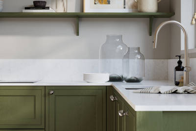 Bewdley Tailored Green painted wood grain Shaker kitchen cabinetry with steel knobs, steel tap, white marbled worktop and matching backsplash, an open shelf on the wall above cabinets with ceramics and a vase of flowers.