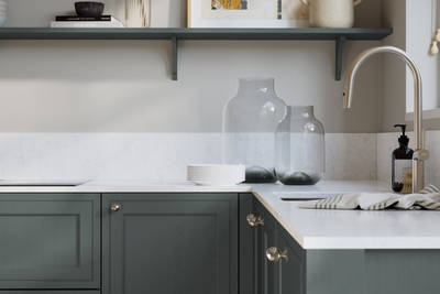 Bewdley Arboretum Green painted wood grain Shaker kitchen cabinetry with steel knobs, steel tap, white marbled worktop and matching backsplash, an open shelf on the wall above cabinets with ceramics and a vase of flowers.