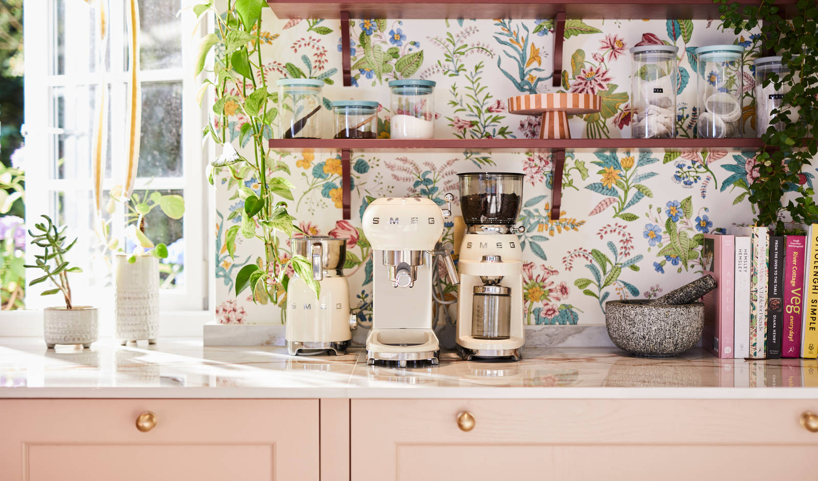 Close-up of Smeg coffee machines on counter top in the coffee area in Sophie Robinson's maximalist kitchen, with shaker style Ludlow Chalk blush cabinetry, open shelving on a wall with a floral wallpaper.