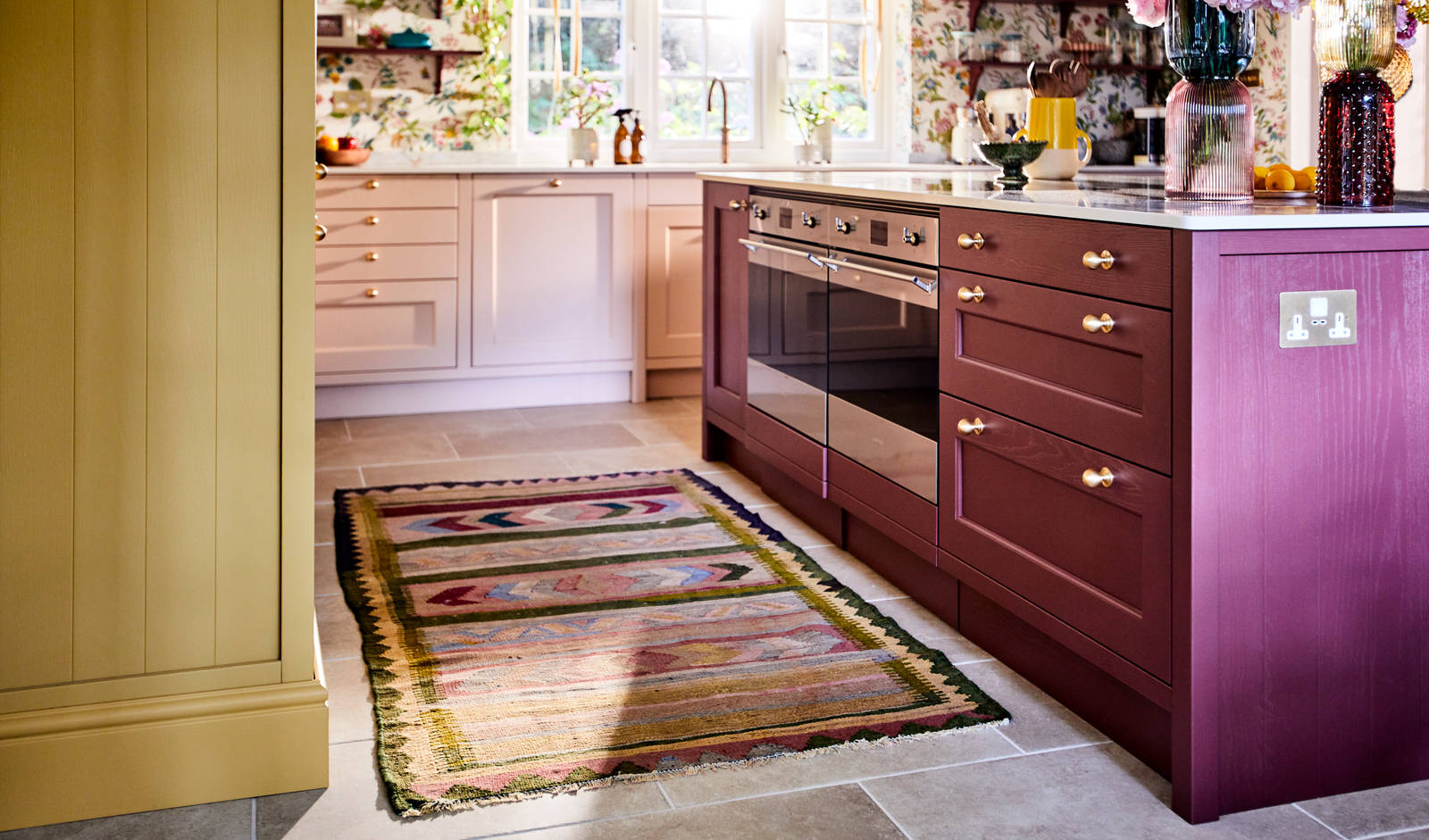 View of Ludlow kitchen island in Burlington Red, ovens in kitchen island and a woven colourful rug on the tile floor.