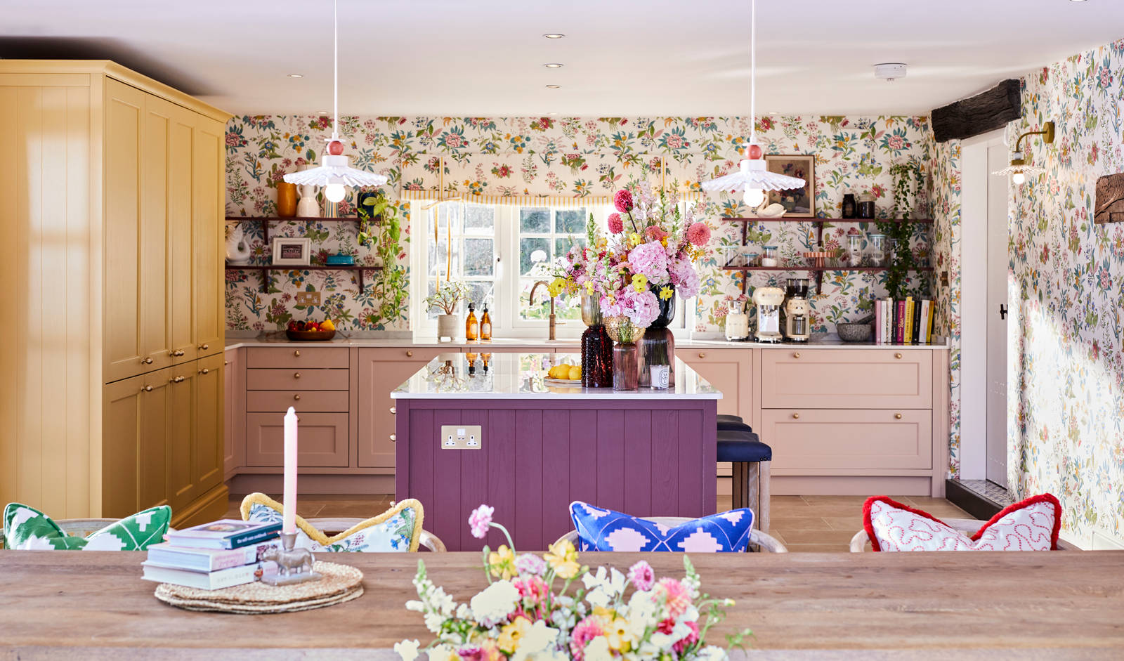 View over wooden table with a pile of books and a candle in foreground and Magnet Ludlow kitchen in background, in Sophie Robinson's vibrant maximalist kitchen.